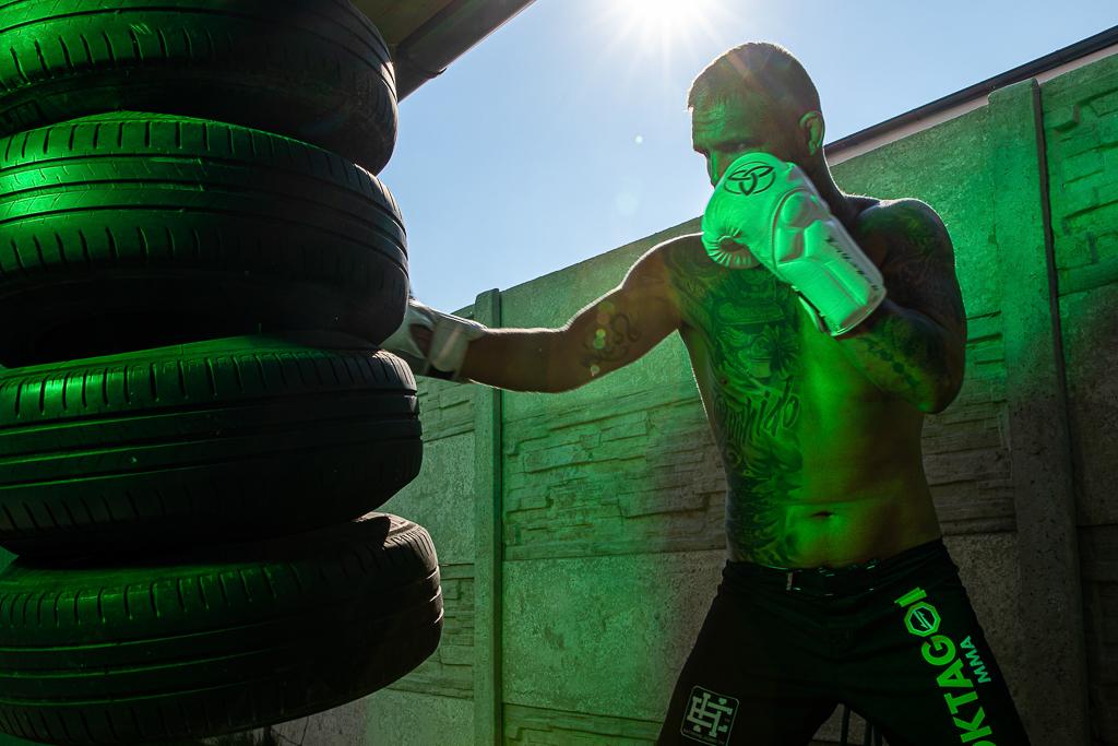 Samuel Krištofič v kraťasech Oktagon, na těle má tetování, na rukou boxovací rukavice a bije do zavěšených pneumatik