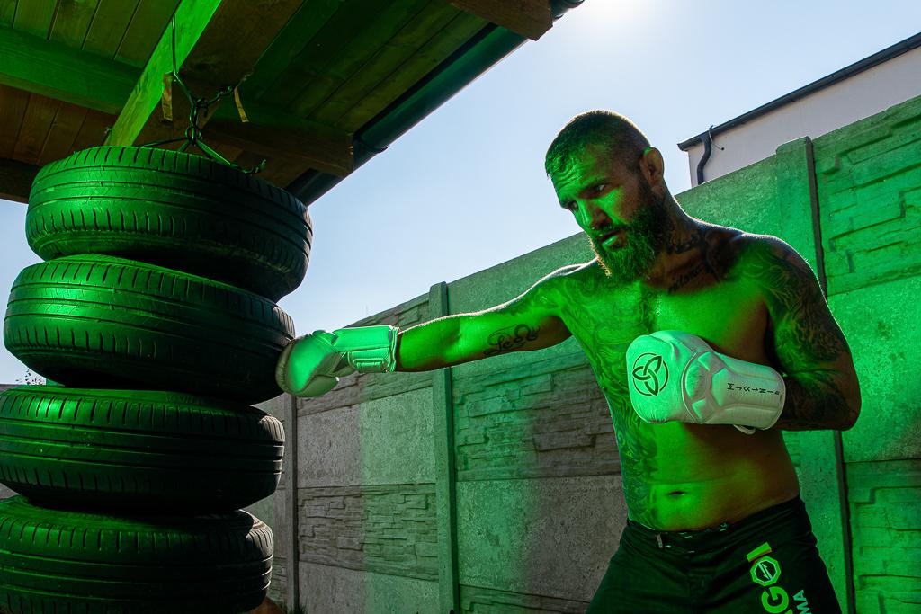 Samuel Krištofič v kraťasech Oktagon, na těle má tetování, na rukou boxovací rukavice a bije do zavěšených pneumatik