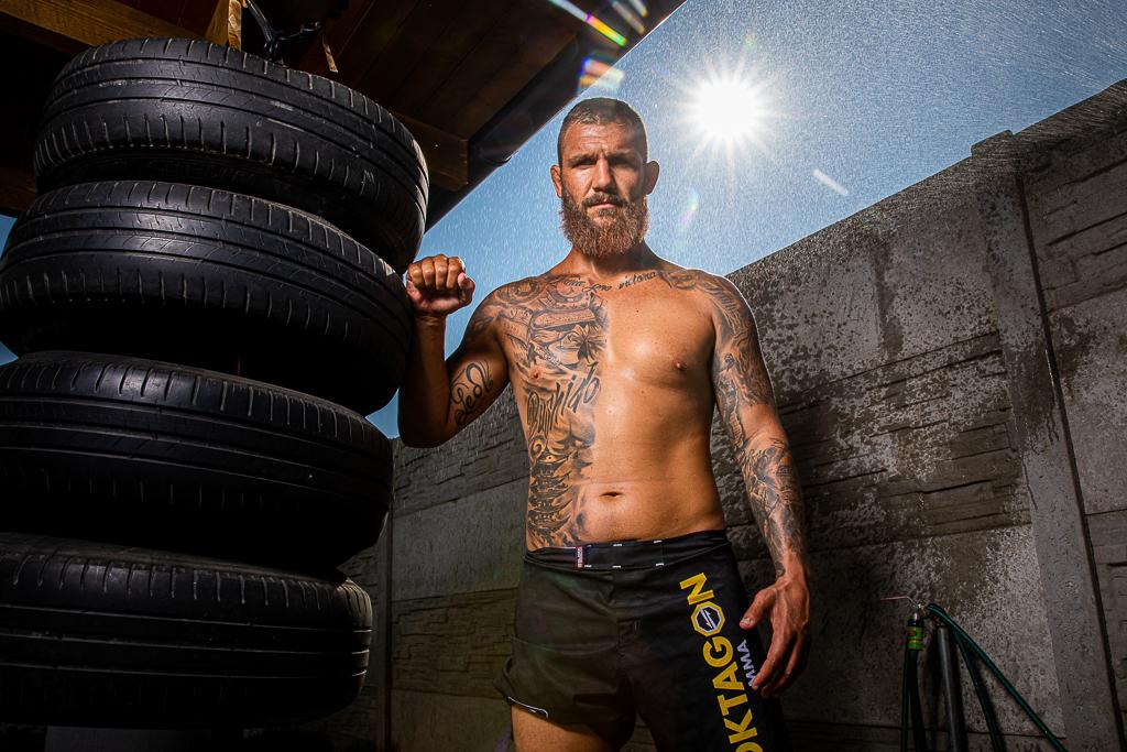 Samuel Krištofič s tetováním na těle, má kraťasy oktagon, stojí vedle zavěšených pneumatik
