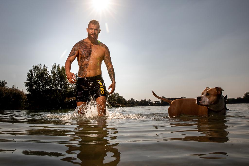 Samuel Krištofič jde ve vodě se psem, na těle má tetování, má kraťasy Oktagon