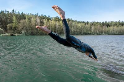 Tomáš Řenč skáče šipku do vody v přírodě, má na sobě dlouhé plavky a plaveckou čepici