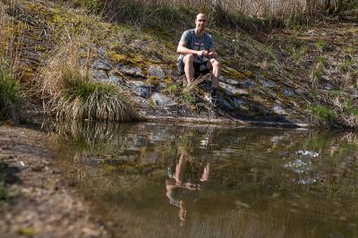 Tomáš Řenč sedí na kamenech u vody v přírodě, ve vodě je jeho odraz