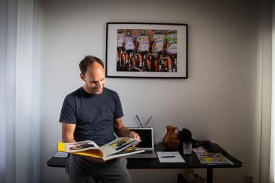 František Raboň si prohlíží cyklistickou knihu , na stole za sebou má notebook, na stěně visí obraz z cyklistického závodu