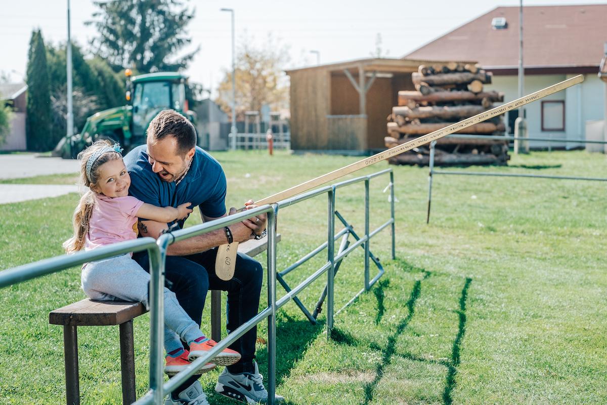 Tomáš Rolinek sedí na lavičce s dcerou u fotbalového hřiště, drží hokejku s podpisy