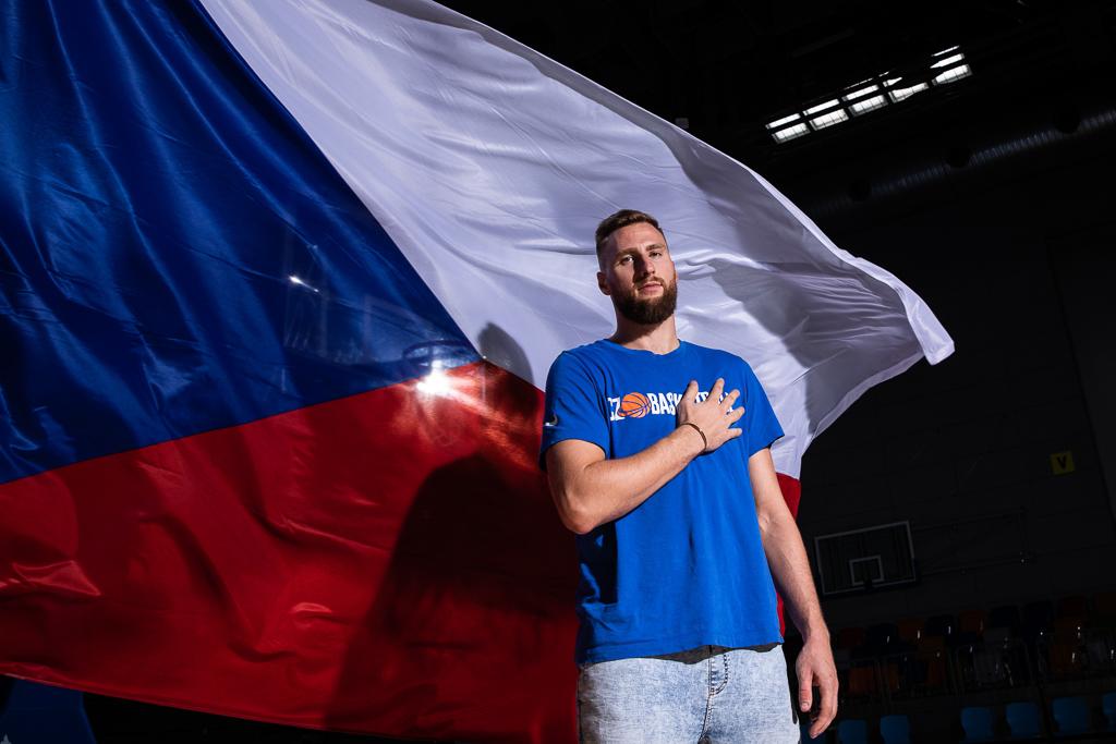 Martin Kříž v tričku českého basketbalu stojí před státní vlajkou a drží se na srdci.