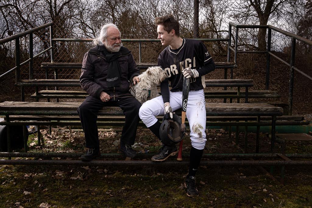 Lukáš Hudeček v baseballovém oblečení s pálkou se svým dědečkem a psem na tribuně