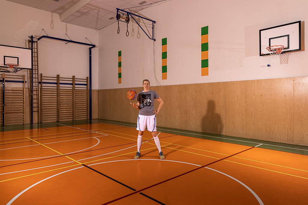 Ivana Večeřová stojí v tělocvičně s basktebalovým míčem, má za sebou basketbalové koše, žebřiny.