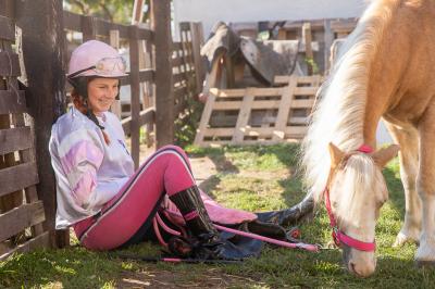 Barbora Miksánková Málková sedí na zemi, opírá se o kůl plotu, má na sobě žokejský oblek a helmu, kouká na koně