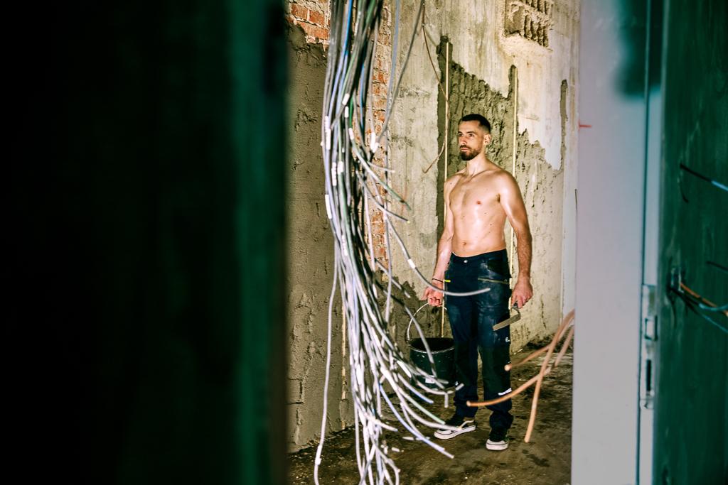 Petr Gewiese bez trička stojí na stavbě, před ním jsou viditelné dráty ze zdi.
