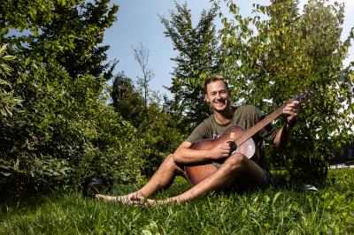 Marek Mazanec sedí v trávě s kytarou, má za sebou stromky