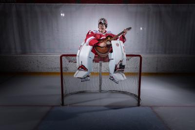 Marek Mazanec v hokejové brankářské výstroji sedí na hokejové brance na ledě, drží v ruce kytaru