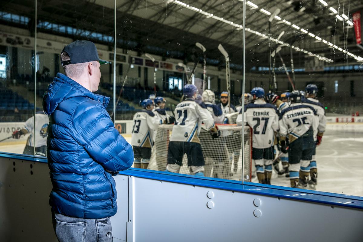 Adam Kubík má založené ruce a dívá se přes sklo na hokejový tým na ledě