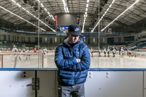 Adam Kubík stojí před hokejovým stadionem, na kterém je vidět hokejový tým, dívá se do země a má založené ruce