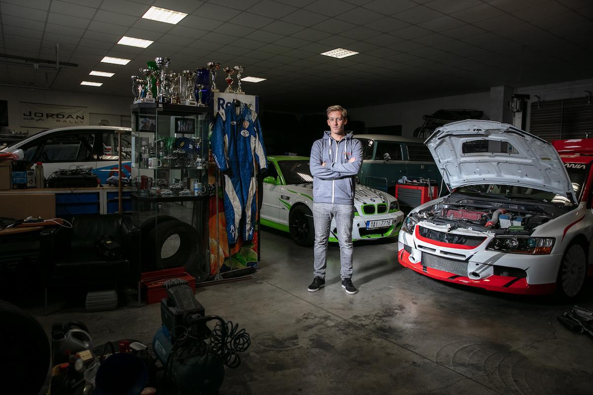 Martin Semerád stojí v garáži a dílně, v pozadí je závodní auto s otevřenou kapotou.